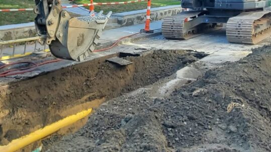 Excavator beside an open street trench with an exposed yellow utility pipe