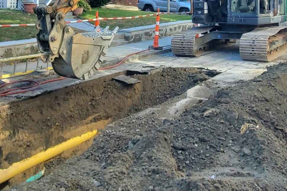 Excavator beside an open street trench with an exposed yellow utility pipe